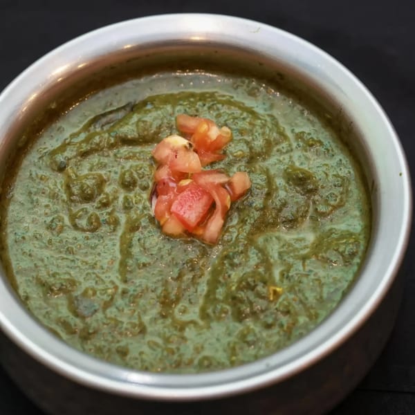 A close-up of green Indian curry with diced tomatoes in a silver bowl.