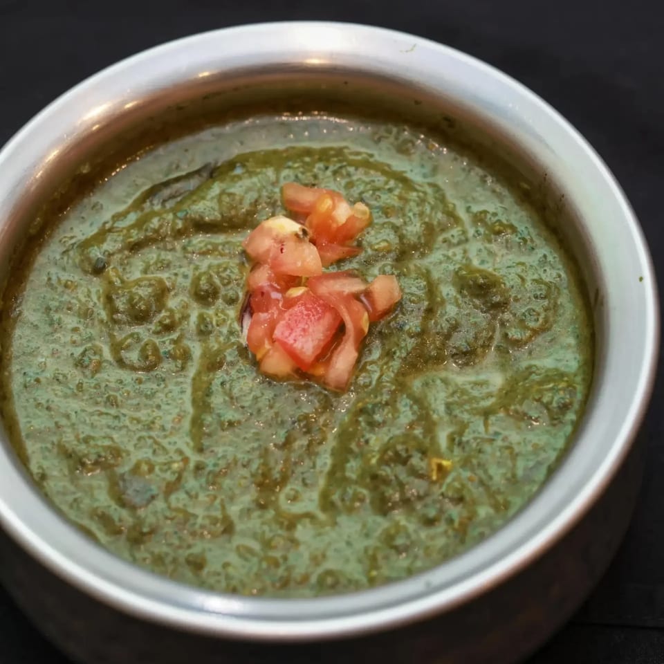 A close-up of green Indian curry with diced tomatoes in a silver bowl.
