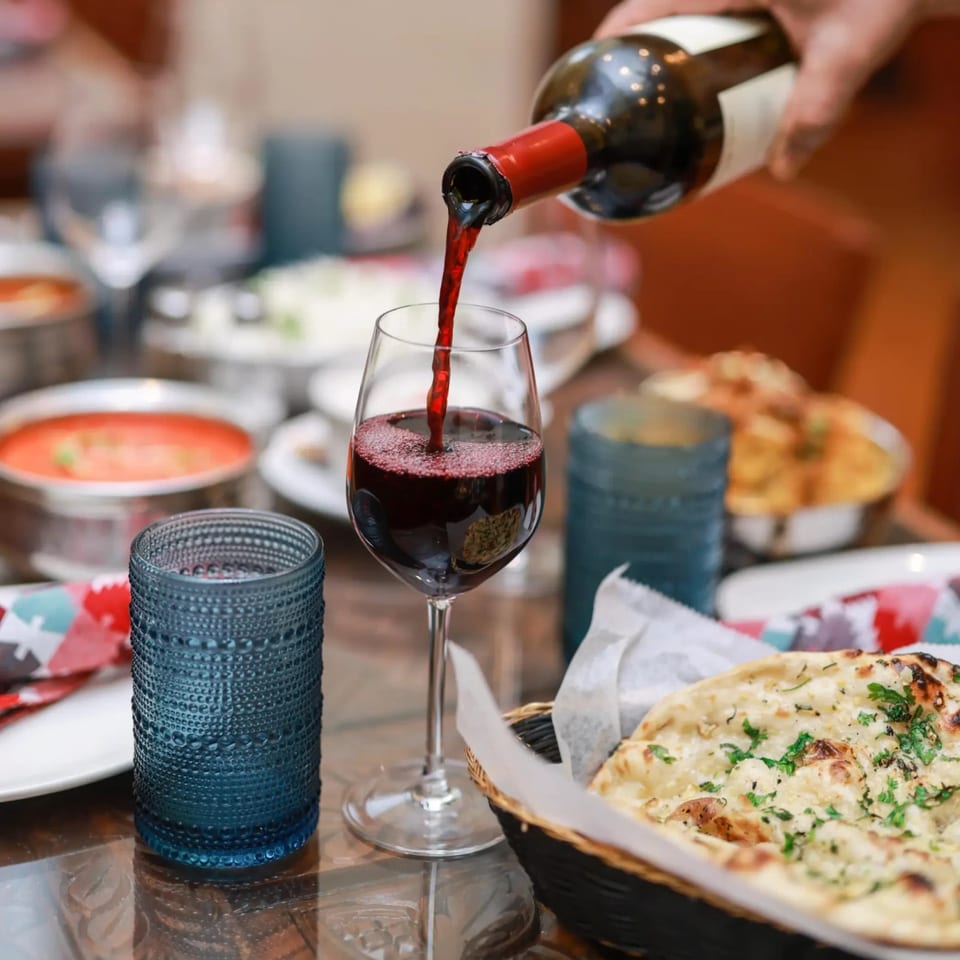 Red wine being poured into a glass at a table with Indian food and naan bread.