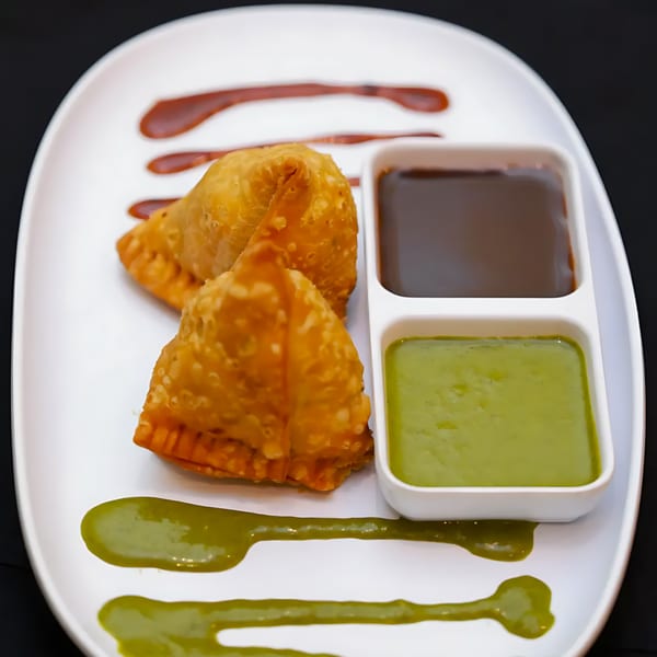 Two golden-brown samosas on a white plate with green and brown dipping sauces.